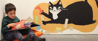 A photograph of a young child sat reading a book on the floor. There is a cartoon illustration of a cat on the wall behind.