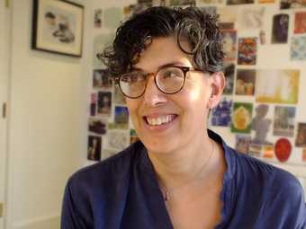 Head-and-shoulders photo of a person smiling, eyes looking away from the camera towards upper left; behind is a wall covered in postcards and images