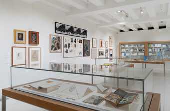 A room with books displayed in 3 glass cases with prints hung on the surrounding walls.