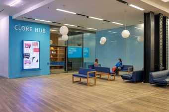 two people sit on sofas outside the Clore hub space. The space has open doors and a digital screen beside it with children's drawings on it and the words 'Tate Kids'